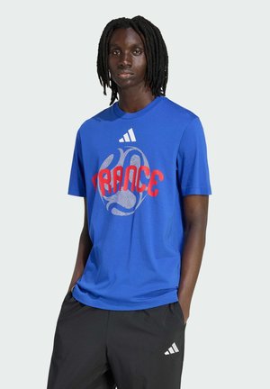 Osoba z dredami ubrana w niebieską koszulkę z czerwonym napisem "FRANCE" i białym logo Adidas, w parze z czarnymi spodniami Adidas.