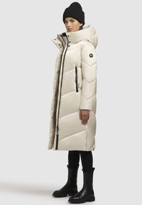 Lang beige pufferjakke med chevron-syning, høj hals og frontlynlås. Sammen med sorte leggings og knæhøje sorte støvler.