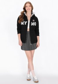 Sudadera negra con texto "MY MO" en blanco en el frente, con capucha fruncida, combinada con una falda plisada gris y zapatillas plateadas.