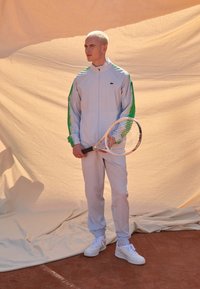 Trening alb de sport cu accente verzi, mâneci texturate și guler cu fermoar. Modelul ține o rachetă de tenis, stând pe o suprafață de zgură.