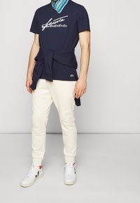 Navy t-shirt with white graphic text, cream joggers, and white sneakers with red accents. A blue patterned bandana is tied around the neck.