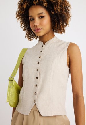 Junge Frau mit lockigem Haar, die ein ärmelloses, beigefarbenes, geknöpftes Oberteil trägt und eine limettengrüne, strukturierte Umhängetasche hält.