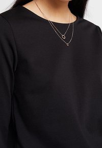 Zwei geschichtete Halsketten aus Roségold; eine dünne Kette mit einem Kreisanhänger und eine andere mit einem länglichen ovalen Akzent. Getragen über einem schwarzen Oberteil.