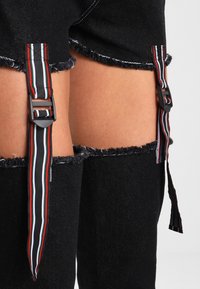 Svart denimshorts med fransade kanter, dubbel spänne med röd, vit och grön rand, samt utskärningar på sidorna.