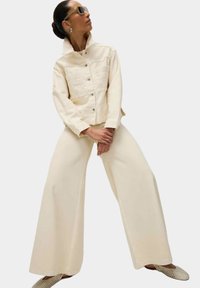 Giacca di jeans crema con bottoni frontali e dettagli in pizzo. Pantaloni a gamba larga crema e scarpe beige texture completano l'outfit. Gli accessori includono occhiali da sole.