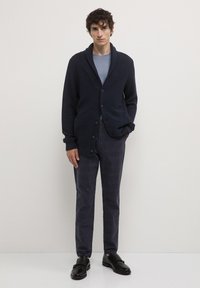Cardigan texturé bleu marine avec col châle, fermeture à boutons, porté sur une chemise bleu clair et un pantalon à carreaux foncés, associé à des chaussures noires.