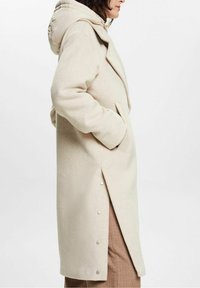 Gabardina beige con capucha, hecha de tela suave. Presenta un corte largo, bolsillos laterales y detalles de botones a lo largo del dobladillo.