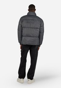 Puffige graue Jacke mit hohem Kragen und gepolstertem Design; mit Branding auf dem Rücken. Kombiniert mit schwarzen Cargo-Hosen und weißen Sneakers.