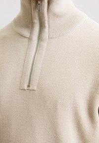 Beżowy sweter z dzianiny z kołnierzem na pół-zamek. Posiada fakturowany materiał, prążkowany dekolt i schludny detal z zamkiem.