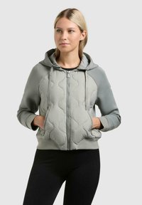 Kvinde iført en lysegrå quiltet hættetrøje med lynlås, snore og lommer, kombineret med sorte tætsiddende bukser, stående foran en ensartet baggrund.
