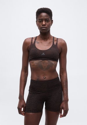 Femme athlétique debout, portant un soutien-gorge de sport sombre à motifs et un short, montrant un tatouage de lion sur le haut de l'abdomen et un nombril percé.