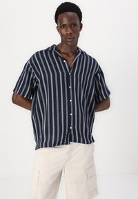 Chemise à manches courtes rayée bleu marine et crème avec col à revers et patte de boutonnage, faite d'un tissu léger, présentée sur fond blanc.