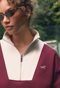 Tumši sarkans džemperis ar rāvējslēdzēju, ar krēmkrāsas apkakli un izšūtu "self." logotipu. Aprīkots ar sudraba rāvējslēdzēja detaļām un mīkstu, teksturētu audumu.