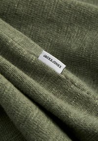 Gros plan d'un tissu tricoté vert olive avec une étiquette blanche portant la mention « JACK & JONES » cousue sur le matériau.