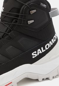 Czarne wodoodporne buty hikingowe z teksturowanym materiałem, białym logo i metalowymi akcesoriami. Posiadają solidną białą podeszwę z rowkami w celu zwiększenia trakcji.