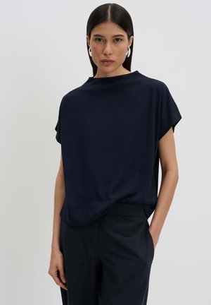 Giovane donna con capelli scuri e lisci che indossa una camicetta blu navy a maniche corte e pantaloni scuri, in piedi con una mano in tasca.