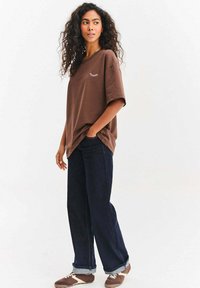 Mladá žena s kudrnatými vlasy oblečená v hnědém oversized tričku, tmavých džínách s širokými nohavicemi a odpovídajících hnědých teniskách, stojící s jednou rukou v kapse.