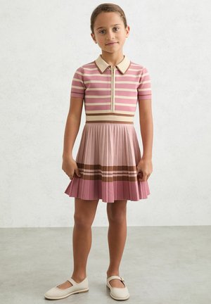 Lyserød og cremefarvet ternet kjole med krave og plisseret nederdel. Har vandrette brune striber nær kanten. Bæres med hvide sko.