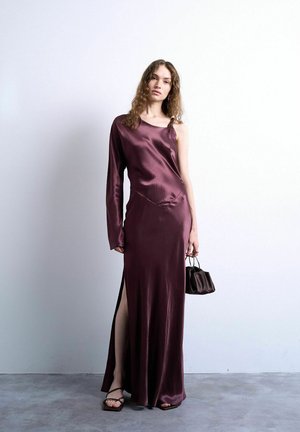 Kvinde iført en gulvlængde, asymmetrisk satin kjole med et langt ærme, der holder en lille mørk håndtaske, stående mod en ensfarvet væg.