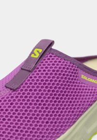 Μωβ παπούτσι τύπου slip-on με ανώτερο μέρος από δίχτυ, υφή με εξαγωνικά σχέδια, μαλακό μωβ λουράκι και κίτρινη λεπτομέρεια λογότυπου.