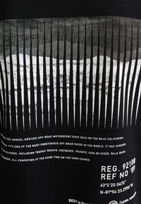 Tessuto nero con testo grafico bianco e linee verticali. Il testo descrive la gara fuoristrada Baja 1000. Include dettagli di registrazione e posizione.