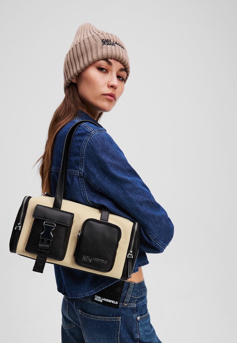 Beige und schwarze Handtasche mit strukturiertem Design, doppelten Reißverschlussfächern und einer Fronttasche mit Schnallendetail. Aus Stoff und Leder.