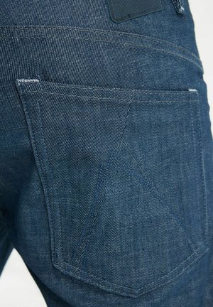 Detalle del bolsillo trasero de unos vaqueros de mezclilla azul, mostrando las costuras y un diseño geométrico sutil en la tela.