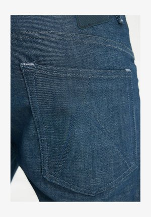 Sinise teksapüksi tagatasku detail, kus on näha õmblusi ja kangal peen geomeetriline mustriline kujundus.