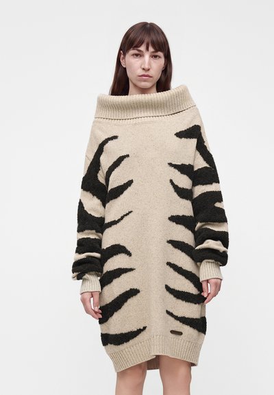 Femme portant une robe-pull beige épaules dénudées avec des rayures noires abstraites, debout devant un fond clair uni.