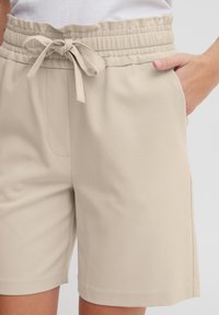 Shorts beiges avec une taille élastique ornée d'un nœud papillon, poches latérales et une texture de tissu lisse. Design classique et décontracté.