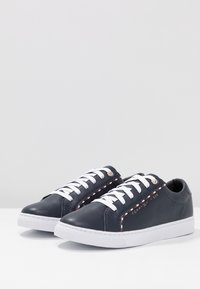 Sneakers in pelle blu navy con lacci bianchi, superficie strutturata e marchio discreto. Presentano un dettaglio a strisce rosse e bianche lungo il lato. Suola in gomma.