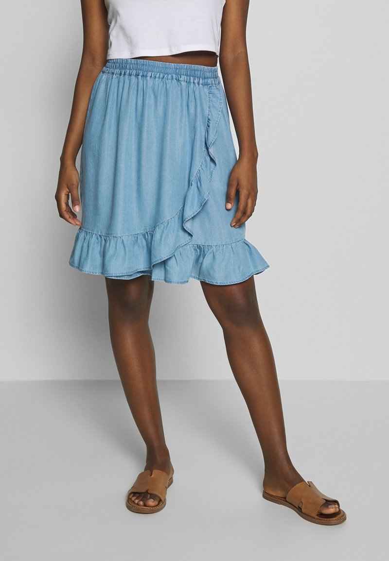 Hellblauer Jeansrock mit fransigem Saum, elastischem Bund und asymmetrischem Wickeldesign. Kombiniert mit braunen flachen Sandalen.