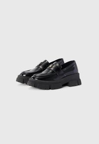 Loafers din piele patent neagră cu talpă din cauciuc masiv, având un superior neted, cusături minime și un detaliu decorativ cu curea.