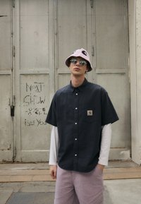 Camicetta a maniche corte con motivo scuro e logo sulla tasca, sovrapposta a una camicia a maniche lunghe bianca. Pantaloni corti viola chiaro e un cappello a secchiello rosa.
