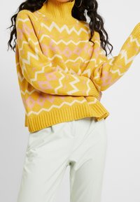 Żółty sweter dziergany z wzorami w zygzak i diamenty w białym i różowym kolorze. Wysoki dekolt i ściągacze na rękawach. Połączony z jasnozielonymi spodniami.