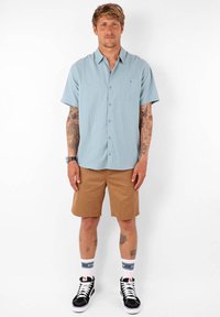 Chemise à manches courtes bleu clair avec une poche, associée à un short beige, des chaussettes blanches avec des accents bleus, et des baskets montantes noires.