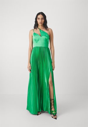 Abito da sera verde in satin con gonna plissettata, scollatura asimmetrica e una spacco alto fino alla coscia. Abbinato a sandali neri con lacci.