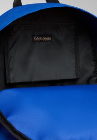 Interior de una mochila azul que presenta un bolsillo negro con cremallera y una etiqueta de marca con el texto "NAPAPIJRI". Fabricada con un tejido duradero y texturizado.