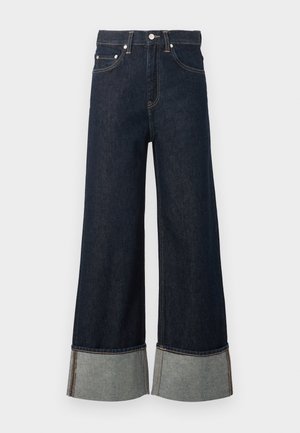 Jean denim bleu foncé à jambes larges avec revers roulés montrant un tissu intérieur plus clair et une fermeture par bouton argenté.