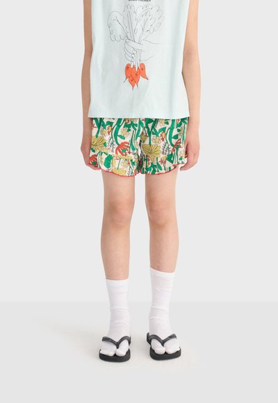 Enfant portant des chaussettes blanches avec des tongs noires, un short à motif floral et un haut sans manches bleu clair avec un dessin de légumes fait à la main.