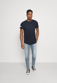 Marineblå kortærmet t-shirt med en hvid detalje på ærmet, parret med lyseblå skinny jeans og sorte sneakers.