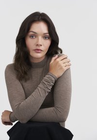 Brown long-sleeve top with a ribbed texture and high neckline, paired with a dark fabric skirt. Gold ring on the model's hand.