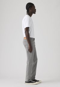 Άνδρας με πλεξούδες στέκεται πλαγίως φορώντας λευκό μπλουζάκι, γκρι τζιν και μαύρα αθλητικά παπούτσια σε απλό φωτεινό φόντο.