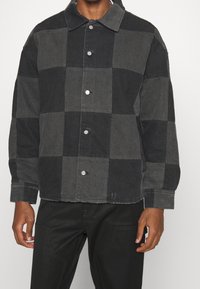 Karierte Jeansjacke in Schwarz und Grau mit silbernen Knöpfen, Kragen und lockerem Schnitt mit sichtbaren Nähten und ausgefranstem Saum.