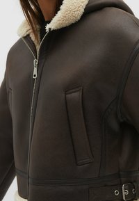 Veste en simili cuir marron avec un col en shearling, fermeture éclair, deux poches latérales et un détail de ceinture en tissu à la taille.