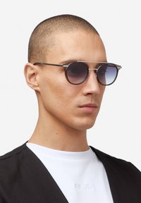 Kulaté sluneční brýle s tmavými gradientními čočkami, kovovým rámem a tenkým mostem nad nosem. Model nosí černou vestu přes bílou košili s logem.