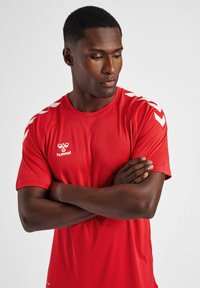 Camiseta deportiva roja de manga corta, con detalles en blanco en los hombros y un logo. Fabricada con material transpirable y elástico.
