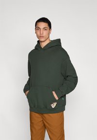 Πράσινο βαμβακερό hoodie με τσέπη καγκουρό και πλευρική λεπτομέρεια εφαρμογής. Το μοντέλο φοράει καφέ παντελόνι και στέκεται μπροστά σε ουδέτερο φόντο.