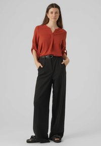 Blusa leggera di color ruggine con scollatura a V e maniche arrotolate, abbinata a pantaloni neri a gamba larga. Insieme, delle infradito nere piatte completano il look.