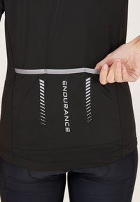 Svart sportig topp med reflexdetaljer, prydd med en silverlogotyp "ENDURANCE" och en liten bakficka, tillverkad av strukturerad, fuktavvisande tyg.